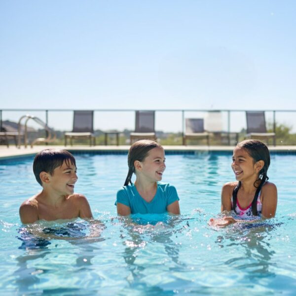 Three diverse children, a boy and two girls, smile and splash together in a sunny outdoor swimming pool.