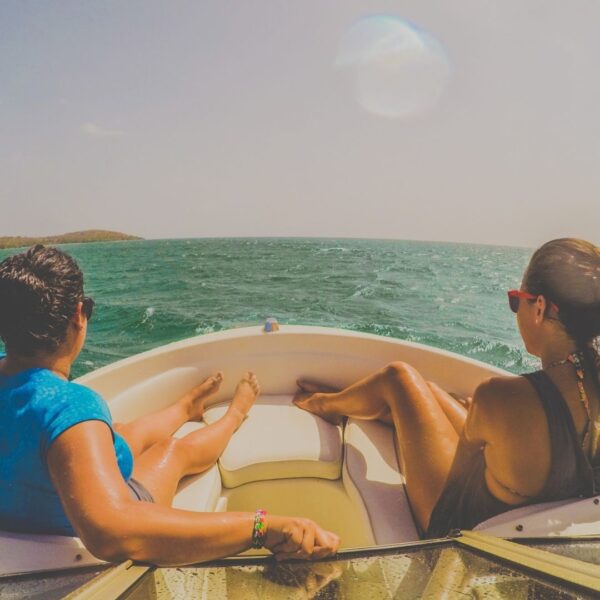 Two people with their backs to the camera sit on a small boat, facing the open, sunny ocean water.